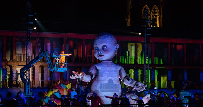 A giant baby model the size of a double decker bus lit up on stage with actors surrounding it