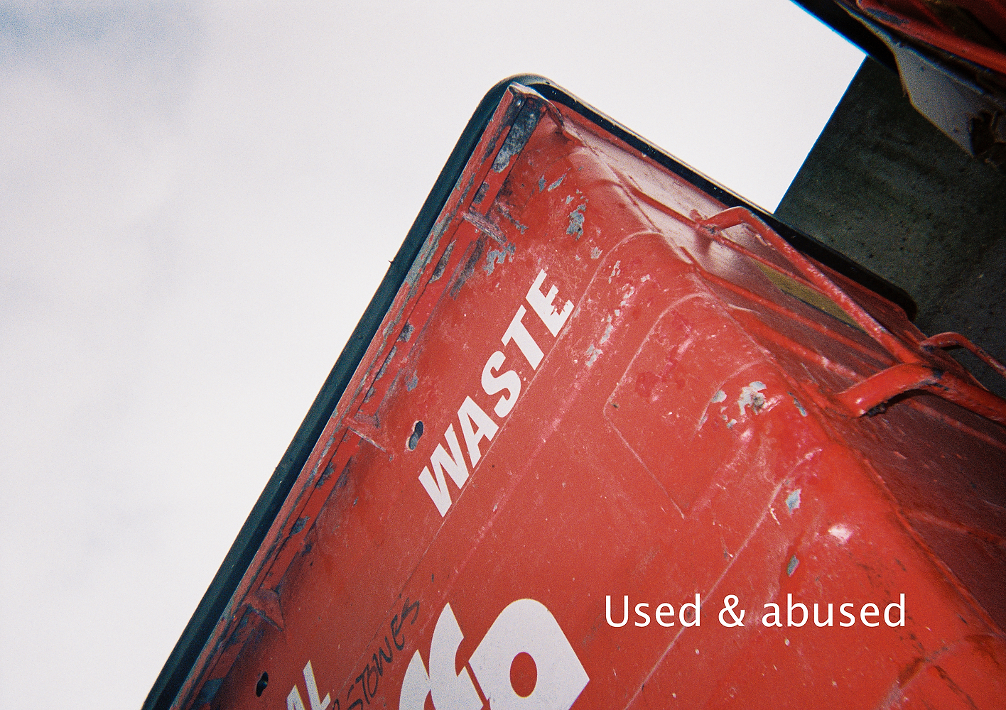 An image of a red industrial rubbish bin taken from a low-angle against a cloudy sky. The bin reads "Waste" and the picture is captioned "Used & abused"
