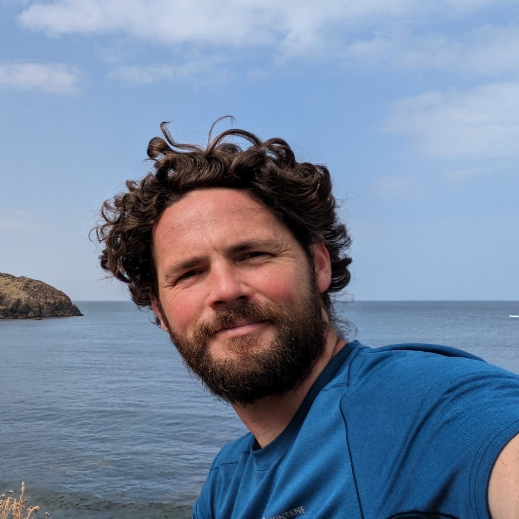 Headshot of John Hallett in a blue t-shirt smiling in front of a coastal backdrop.